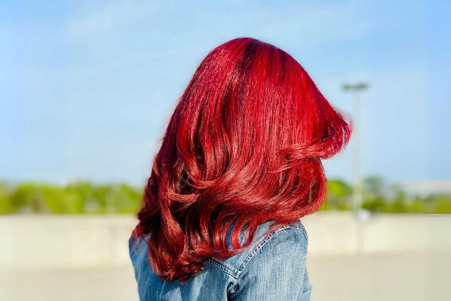 Person with vibrant red hair stands outdoors, facing away, wearing a denim jacket.